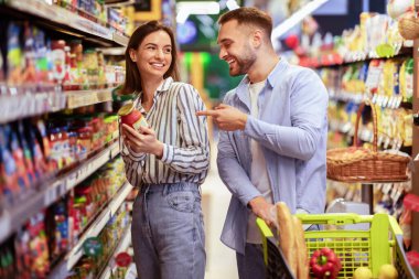Market alışverişi. Genç, gülümseyen bir çift marketten alışveriş yapıyor. İki Alıcı Alışveriş Arabasıyla Koridorda Duruyor Yiyecek Ürünlerini Seçiyor ve Gülüyor