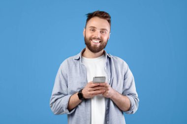 Mavi stüdyo arka planında cep telefonu kullanan havalı sakallı adamın portresi. Modern aletli neşeli genç adam interneti tarıyor, e-postaları kontrol ediyor, sosyal medyada paylaşıyor.