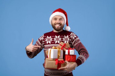Mavi stüdyo arka planında Noel Baba şapkası ve sıcak süveter giyen neşeli genç adamın portresi. Yeni yıl kutlaması için hediyeleri olan mutlu bir milenyum adamı.