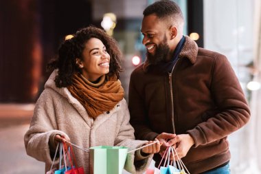 Keyifli alışverişler. Renkli alışveriş torbaları taşıyan mutlu siyah bir çiftin portresi. Yeni kıyafetleri ya da hediyeleri için heyecanlılar. Akşam alışveriş merkezinin yakınında durup birbirlerine bakıyorlar.