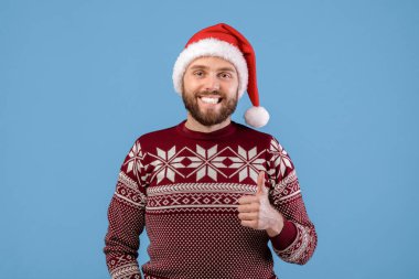 Noel süveteri ve Noel Baba şapkası giymiş pozitif bir genç. Başparmağını gösteriyor, gülümsüyor ve mavi stüdyo arka planındaki kameraya bakıyor. Yılbaşı indirimini ya da özel teklifi destekleyen milenyum adamı