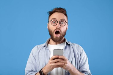 Şok olmuş sakallı adam mavi stüdyonun arka planında cep telefonuna bakıyor. Akıllı telefondan inanılmaz haberler öğrenen şaşırtıcı genç adam internette büyük bir satış yaptı.