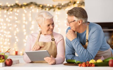 Aged Couple With Tablet Ordering Food Online In Restaurant For Christmas Dinner Sitting In Modern Kitchen At Home. Retired Life, Family Holidays And Cooking Hobby. Recipes And Grocery Shopping App