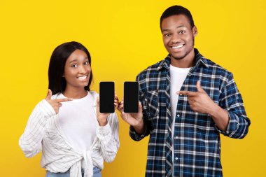 Satın alma ve mağazalardan gelen mesajlar için çevrimiçi uygulama. Gülümseyen zoomerlar Afro-Amerikan çiftler boş ekranlı akıllı telefonlar gösteriyorlar ve sarı arka planda izole edilmiş cihazları işaret ediyorlar.