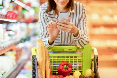 Perakende ve Teknoloji Konsepti. Yakın plan, alışveriş yapan, akıllı telefonunu kullanan, tramvaya yaslanan gülümseyen genç bir kadının görüntüsü. İnternette kadın müşteri kontrolü ve karşılaştırma fiyatları