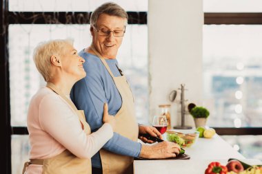 Neşeli yaşlı bir çift birlikte yemek pişiriyor. Evde mutfakta akşam yemeği için salata yapıyorlar. Kıdemli eşler yemek hazırlıyor sebzeleri kesiyor ve gülümsüyor. Metin için Yan Görünüm, Boş Boşluk