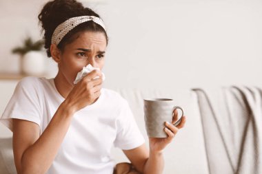 Genç bir kadın kendini hasta hissediyor, burnunu hapşırtıyor, elinde sıcak içecek ve peçeteyle fincanı tutuyor, ev içi fotokopi odasına bakıyor. Esmer bayanın burnu akıyor, üşütmüş ya da koronavirüs enfeksiyonu olmuş.