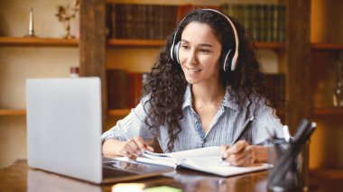 Yetişkinler için öz eğitim. Güzel, pozitif bir genç kadının portresi, kablosuz kulaklık takıyor, online kurs izliyor, masada oturuyor, kişisel bilgisayarını kullanıyor ve not defterine not alıyor.