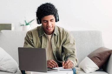 Web tabanlı eğitim konsepti. Bilgisayarda konferans sırasında kulaklık takmış, not defterine yazı yazan genç bir öğrenci. Havalı Afrikalı Amerikalı ev ödevi yapıyor ve Webinar, Panorama dinliyor.