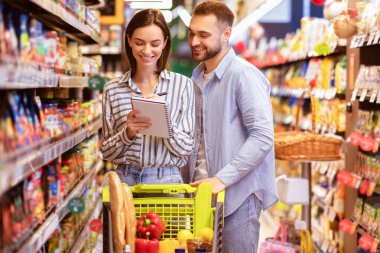 Market alışveriş listesindeki iki mutlu müşterinin portresi, not defterine yazma, ürün satın alma, süpermarkette tam market arabası ile ayakta durma. Genç Milenyum Ailesi Sağlıklı Yemek Seçiyor