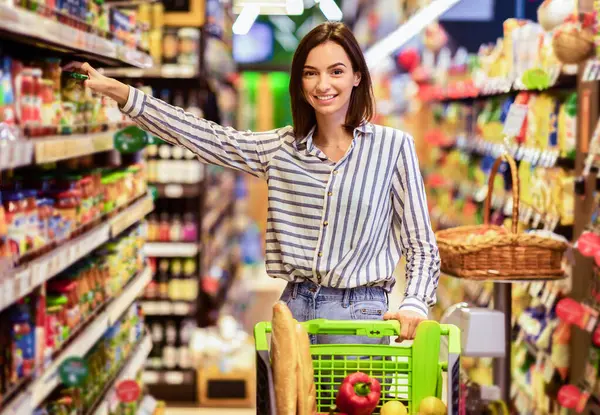 Süpermarkette. Markette Koridorlar Arasında Tramvay Arabasıyla Duran Gülümseyen Genç Kadın. Neşeli Tüketici Süper Market 'te Temelleri Alırken, Raftan Kameraya Bakarken Ürünler Alırken
