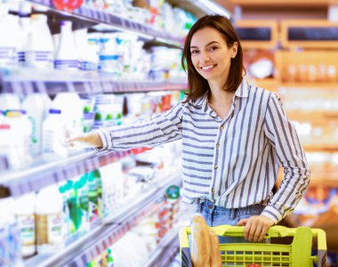 Perakende konsepti. Gülümseyen genç bayan markette rafların yanında alışveriş arabasıyla dikiliyor. Neşeli Kadın, markette temel şeyleri alıyor, mandıra ürünlerini alıp kameraya bakıyor.