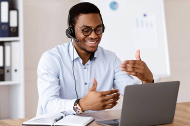 Pozitif Afro-Amerikalı adam ofiste online iş konferansı yapıyor, dizüstü bilgisayar ve kulaklık kullanıyor, müşteriyle ya da iş ortağıyla neşeli bir şekilde konuşuyor, el kol hareketi yapıyor ve gülümsüyor, boşluğu kopyalıyor.