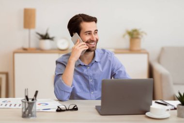 Ofiste telefonla konuşan neşeli iş adamı dizüstü bilgisayarda oturuyor ve çalışıyor. İş yerinde cep telefonuyla konuşan mutlu adam. İş Çağrısı, Mobil İletişim Konsepti