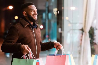 Alışveriş merkezinin vitrininde moda kıyafetlerine bakan gülümseyen Afro-Amerikan bir adamın portresi, akşamları alışveriş merkezinin yakınında yürüyor. Kış Hediyeleri, Satış ve İndirim, Perakende, Satın Alım