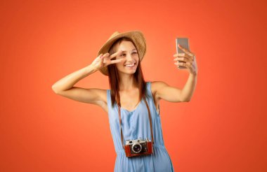 Güzel genç bayan gezgin cep telefonuyla selfie çekiyor, sarı stüdyo geçmişi var. Yazlık şapkalı neşeli kadın sosyal medya için fotoğraf çekiyor, akıllı telefon kullanıyor, el kol hareketi yapıyor.