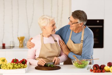 Mutfakta Kıdemli Çiftler Mutfağı. Evdeki masanın yanında duran ve akşam yemeği için sebze keserken karısına sarılan yaşlı bir koca. Yaşlı eşler birlikte yemek hazırlığının keyfini çıkarıyorlar.