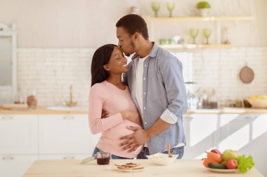 Yakışıklı siyah adam güzel hamile karısına sarılıyor ve mutfakta onu alnından öpüyor. Güzel Afro-Amerikan çifti evde kahvaltı hazırlarken birbirlerini okşuyor.