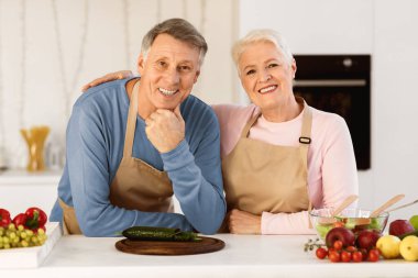 Mutfakta kıdemli çift pozu, evde birlikte yemek pişirip kameraya bakarken masanın yanında dikilmek. Yaşlı Eşler Sağlıklı Yemek Hazırlıyor. Yaşlılar için Beslenme ve Tarifler