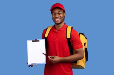 Paket alınıyor. Gülümseyen Afrikalı Amerikalı Sakallı Teslimatçı elinde kalem ve klasörle mal ve yiyecek dağıtıyor, termo çanta giyiyor ve Mavi Stüdyo 'nun arka planında duruyor. Model