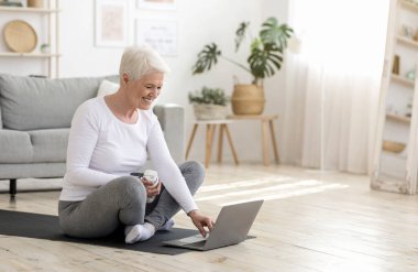 Evde eğitim için internetten özel ders seçen, oturma odasındaki yoga minderinde oturan mutlu yaşlı kadın.
