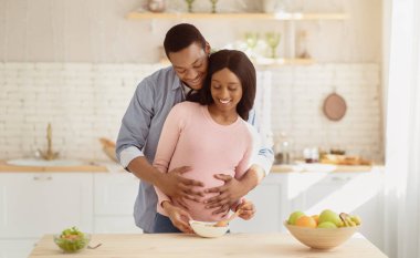 Güzel siyah hamile kadın mutfakta sağlıklı kahvaltı hazırlıyor ve erkek arkadaşı ona arkadan sarılıyor, panorama. Bekleyen güzel çift sabahları evde dengeli yemek yapıyor.