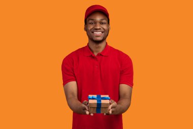 Tatil için teslimat servisi. Gülümseyen Afro-Amerikan kuryenin portresi paketlenmiş bir hediye kutusu tutuyor, kameraya hediye veriyor ve gösteriyor, turuncu stüdyo arka planında izole edilmiş.