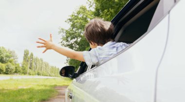 Arabanın camından sarkan küçük çocuğun arka görüntüsü ve uzanmış elleriyle rüzgarı yakalamaya çalışan küçük erkek çocuk yaz havasının ve araba yolculuğunun tadını çıkarıyor, hareket halinde, yakın çekim.