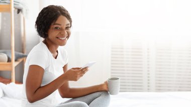 Evdeki yatakta oturan neşeli siyah kadın, cep telefonu kullanıyor, çay içiyor, fotokopi çekiyor. Afrika kökenli Amerikalı bir kadın hafta sonunu evde kahve içip arkadaşlarıyla sohbet ederek geçiriyor.