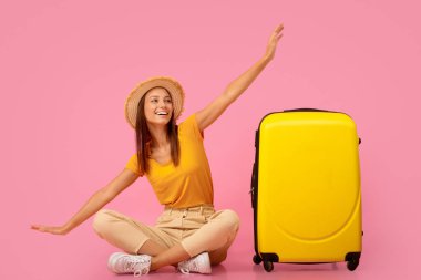Şapkalı mutlu kız bavulun yanında oturuyor, uçağı taklit ediyor, tatili için heyecanlı, pembe stüdyo geçmişi var.