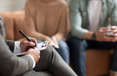 Help Concept. Closeup Cropped View Of Consultant Writing Taking Notes, Selective Focus On Hand With Pen. Spouses Having Session With Male Marriage Therapist, Sitting On Couch In Office.