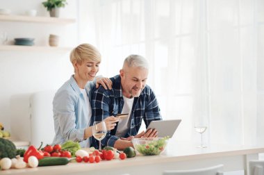 Orta yaşlarda sağlıklı beslenmek, faturaları ödemek, alışveriş yapmak ve internetten alışveriş yapmak. Gülümseyen yaşlı çift modern konfor mutfağında parlak renkli sebzelerle dizüstü bilgisayar ve kredi kartı kullanıyor.