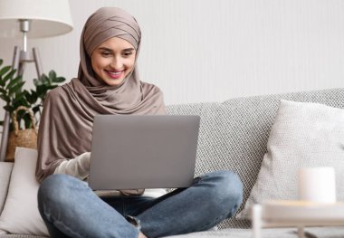 Booking vacation online. Portrait of young muslim woman in hijab using laptop, making hotel and tickets reservation while sitting on couch at home