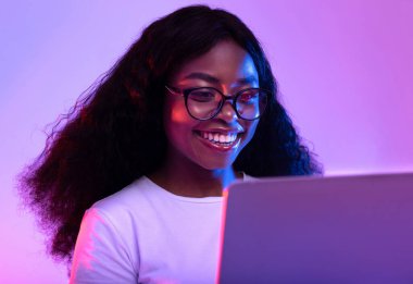 Gözlüklü mutlu siyahi kadın bilgisayarını internet işi için kullanıyor ya da neon ışıkta çalışıyor. Neşeli Afrikalı Amerikalı kadın uzaktan video konferansı yapıyor, arkadaşlarıyla internette sohbet ediyor.