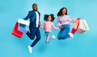 Birlikte alışveriş yapmak. Havada zıplayan, Turkuaz Stüdyo 'nun arka planında izole edilmiş bir sürü parlak alışveriş torbası taşıyan heyecanlı üç Afrikalı Amerikalı ailenin el ele tutuşması.