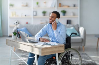 Evden bilgisayarda çalışırken tekerlekli sandalyedeki neşeli, engelli siyahi adam telefonla konuşuyor. Genç engelli iş adamı cep telefonuyla müşteriyle konuşuyor.