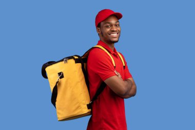 Teslimat Servisi. Kırmızı üniformalı ve termo çantalı özgüvenli siyah erkek kurye portresi katlanmış kollarla poz veriyor ve mavi stüdyo arka planında izole edilmiş kameraya bakıyor.
