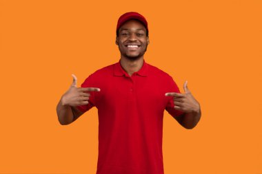 Beni seç. Gülümseyen sakallı Afrikalı Amerikalı teslimatçı kendini işaret ediyor ve kırmızı tişörtlü reklamcı sarı arka planda kurye servisi pozu veriyor. Stüdyo Çekimi