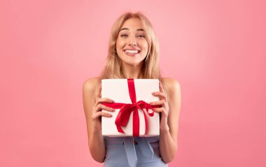 Bahar tatili kutlaması. Güzel elbiseli neşeli bin yıllık bayan, kadınlar günü için hediye paketi ya da pembe arka planda doğum günü kutusunu gösteriyor. Genç sarışın özel bir sürprizle mutlu oluyor.
