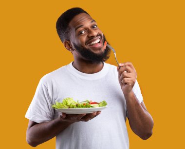 Neşeli siyahi adam salata yiyerek tabağı tutuyor ve çatalla sağlıklı yemeğin tadını çıkarıyor sarı stüdyonun arka planında dikiliyor kameraya gülümsüyor. Vejetaryen yemek tarifleri, kilo kaybı ve erkek beslenmesi.