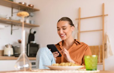 Yemek yerken, masada otururken ve telefonda mesajlaşırken, sosyal ağlara göz atarken ya da online alışveriş yaparken, mutfakta akıllı telefon kullanan heyecanlı bir bayan.