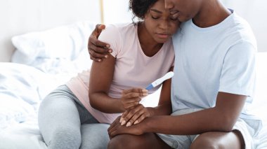 Negatif gebelik testi yapan üzgün siyah çift, depresyondaki karısına sarılan koca, yatak odası içi, boş alan. Üzgün Afro-Amerikan ailesi hamilelik testine bakıyor.