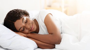 Sağlıklı uyku, uyku hijyeni konsepti. Evdeki yatağında uyuyan, gülümseyen, genç, siyah bir kadın. Afro-Amerikalı kadın uyuyor, beyaz, rahat yatak odasında dinleniyor.