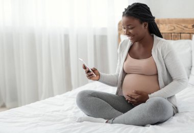 Gülümseyen siyah hamile kadın evde yatağında akıllı telefonuyla oturuyor, heyecanla cep telefonu ekranına bakıyor, mutlu Afrikalı kadın gebelik uygulaması ya da cep telefonu teklifi alıyor,