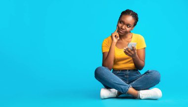 Mavi Stüdyo Arkaplanı Üzerinde Oturan Cep Telefonu Kullanan Düşünceli Afrikalı Amerikalı Kız. Mobil Uygulama Konsepti. Boşluklu Panorama