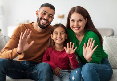 Hoş geldiniz. Evdeki koltukta oturmuş kameraya gülümseyen üç kişilik neşeli Orta Doğulu bir aile. Merhaba, Jest Konsepti. Video Çağrısı ve Modern Uzaktan İletişim