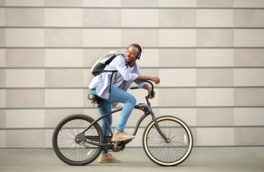 Yakışıklı siyah adam bisiklet sürüyor ve dışarıda tuğla duvarın yanında müzik dinliyor.