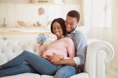 Evdeki kanepede hamile karısına bebek ayaklarıyla sarılan siyahi bir adam. Şirin ayakkabıları olan romantik Afrikalı Amerikalı çift kanepede birbirlerine sarılıp aile olarak huzurlu anların tadını çıkarıyorlar.