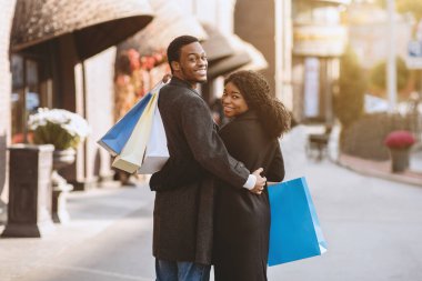 Sonbaharda ve mevsimlik indirimli karaborsa cuma indirimi. Milenyum Afrikalı Amerikalı erkek ve kadın eğleniyor, sarılıyor, renkli kağıt torbaları tutuyor ve alışveriş merkezinin yakınındaki kameraya bakıyor.