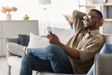 Yeni Mobil Uygulama. Neşeli siyah adam cep telefonu kullanıyor, gözlük takıyor, mesaj atıyor ve evdeki koltukta oturup internete bakıyor. İçerde Kullanıcı Kullanımı Uygulaması. Yan Görünüm